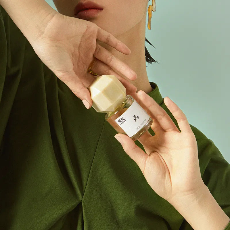 A hand holding a TO SUMMER Triple Tea perfume bottle against a deep green backdrop