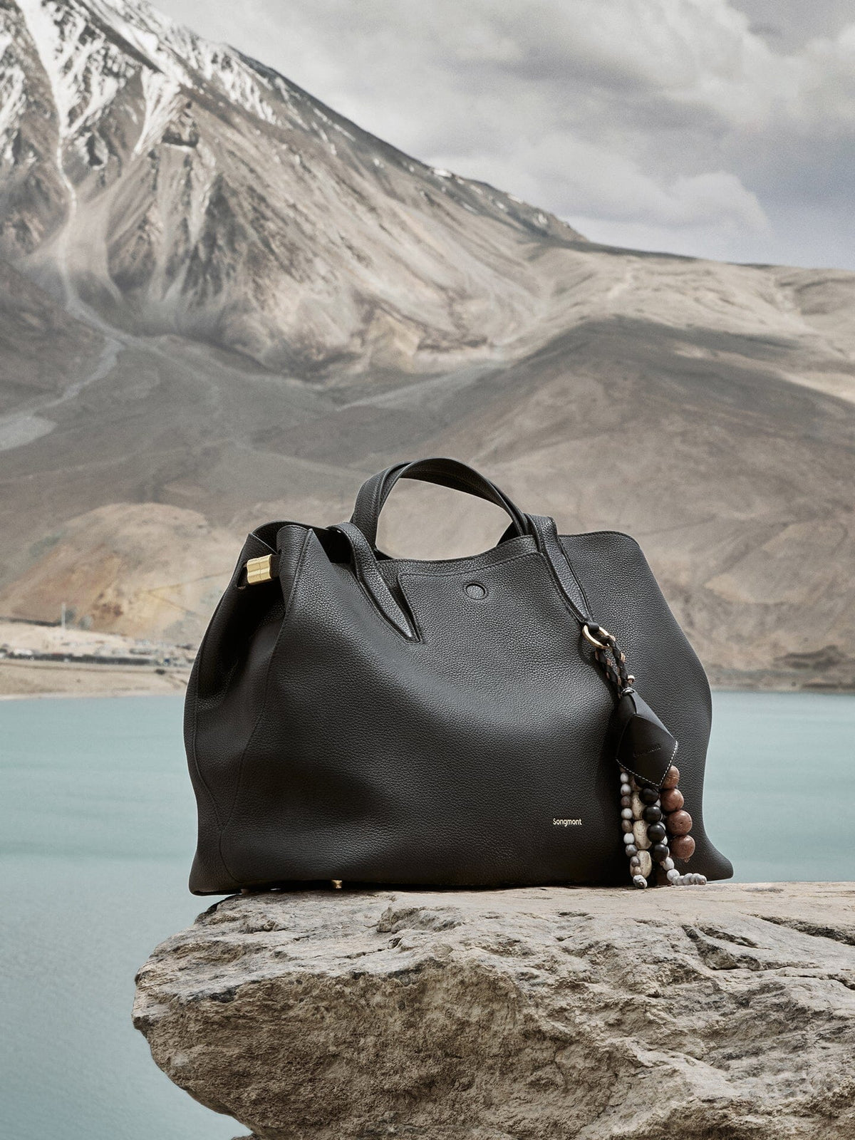 Songmont Large Yore Tote Bag Onyx - on a rock by a mountain lake with a beaded charm