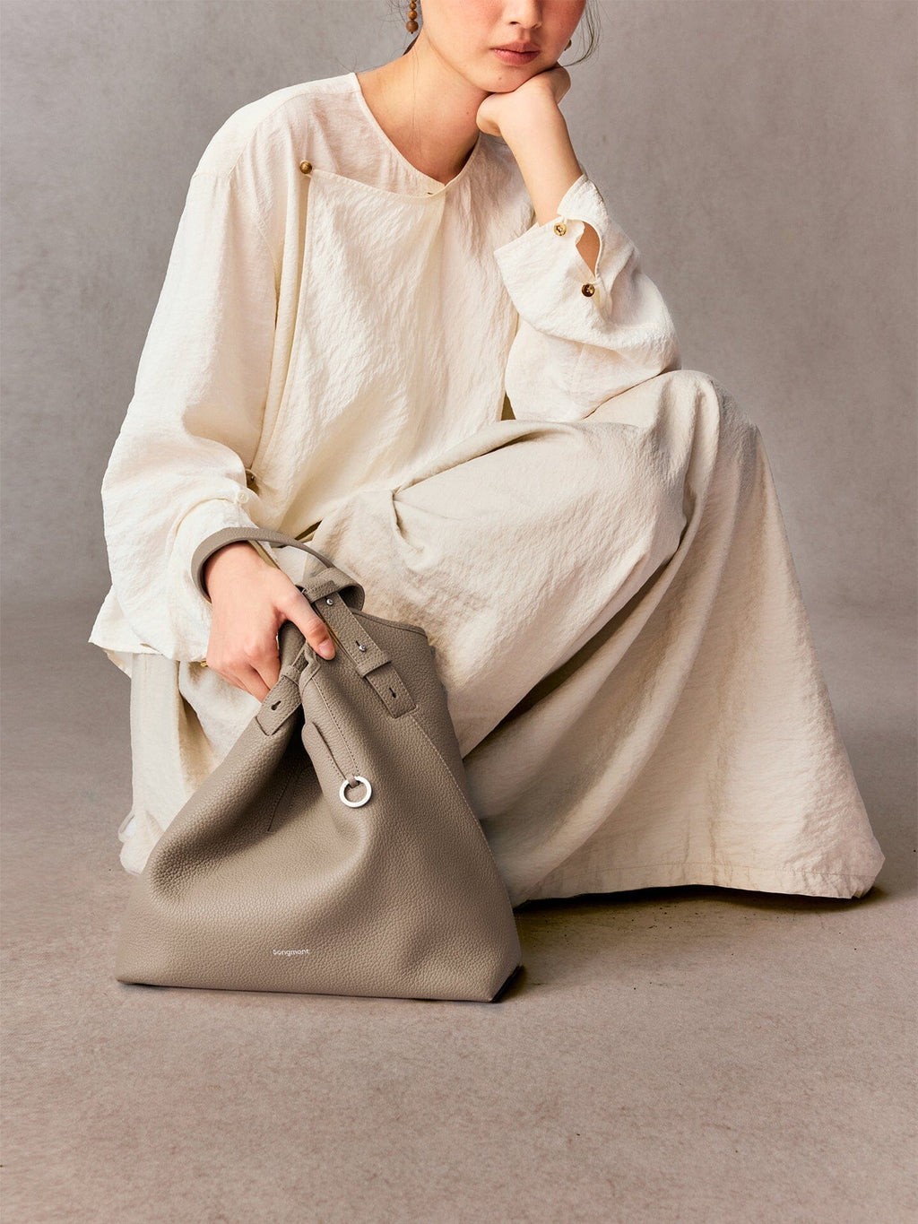 Songmont Medium Yore Shoulder Bag Smoke Grey - held by the strap beside a seated model